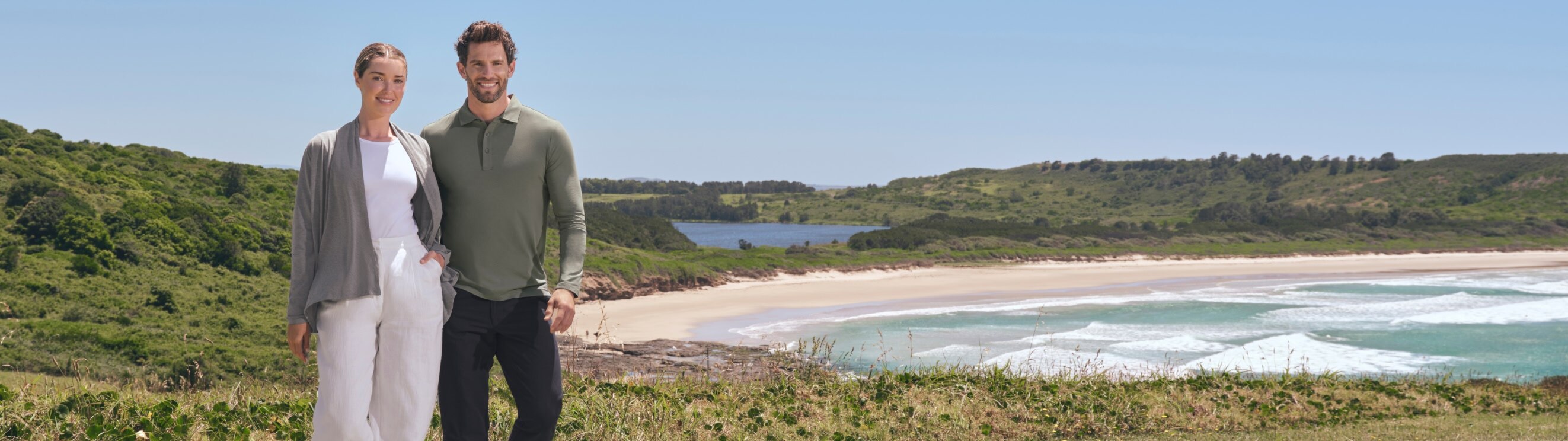 Man and woman standing on a grassy area with a scenic beach and mountains in the background.