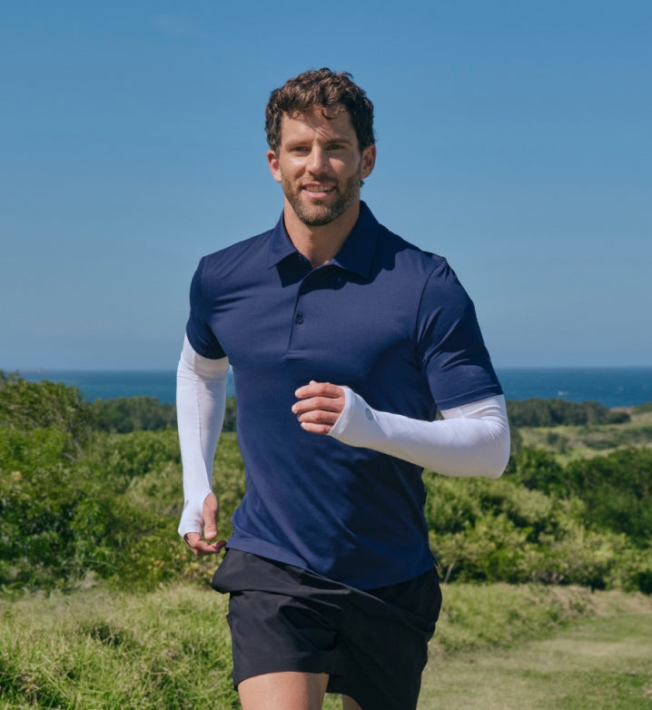 Man running outdoors with UPF 50+ white arm sleeves, a blue polo shirt and black shorts against a scenic background.
