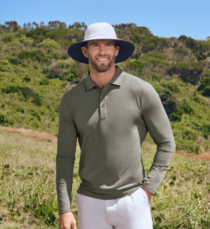 Man wearing a green long-sleeve polo shirt and white pants standing in a grassy field with a blue sky, wearing a Solbari sun hat and sun protective clothing.