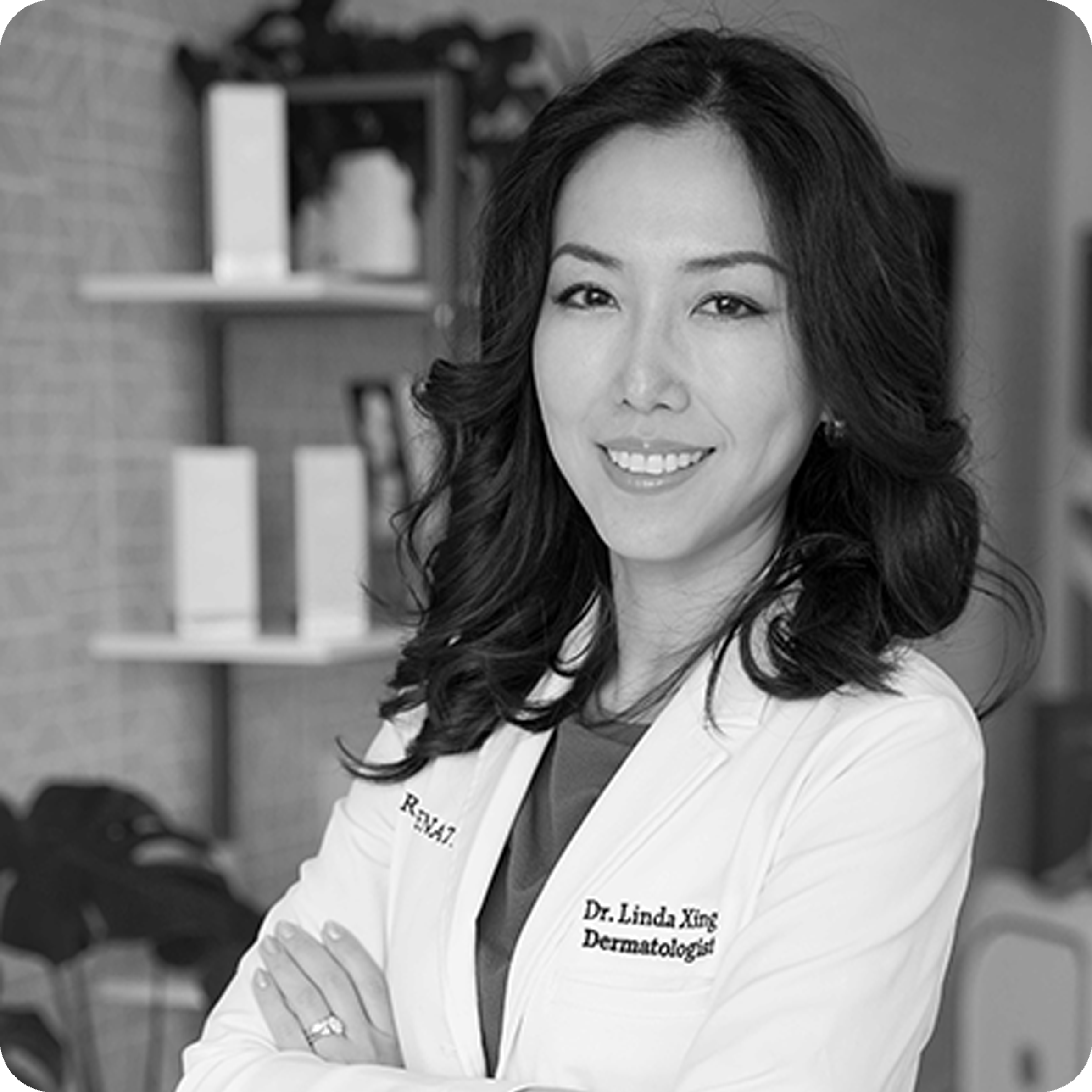 Black and white photo of a woman in a white lab coat with 'Dermatologist' on it, standing in an office setting.
